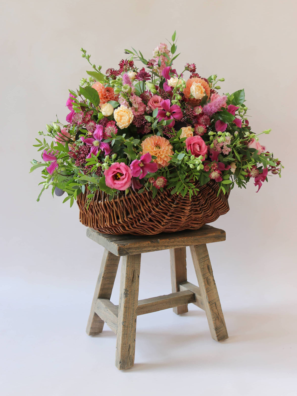 Arrangement of bright pink and peach flowers with green foliage in a wicker basket on a wooden stool against a light background, photographed at a slight angle.