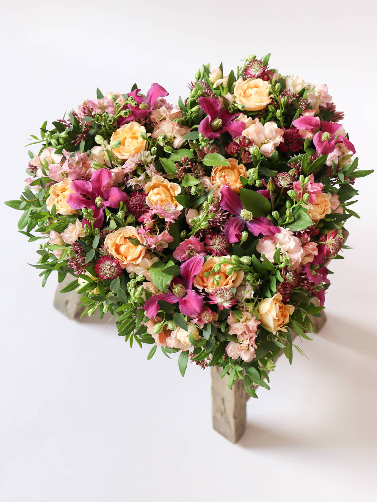 Heart-shaped funeral posy with bright pink and peach flowers on a white background sitting on top of a wooden stool.