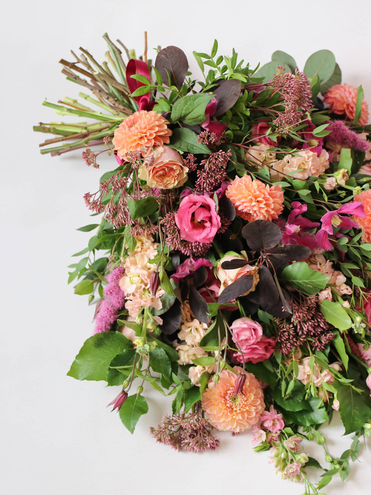 Portrait photograph of a tied funeral sheaf with flowers including pink Lisianthus, peach Stocks, pink Delphiniums and peach Dahlias, tied with a burgundy satin bow.