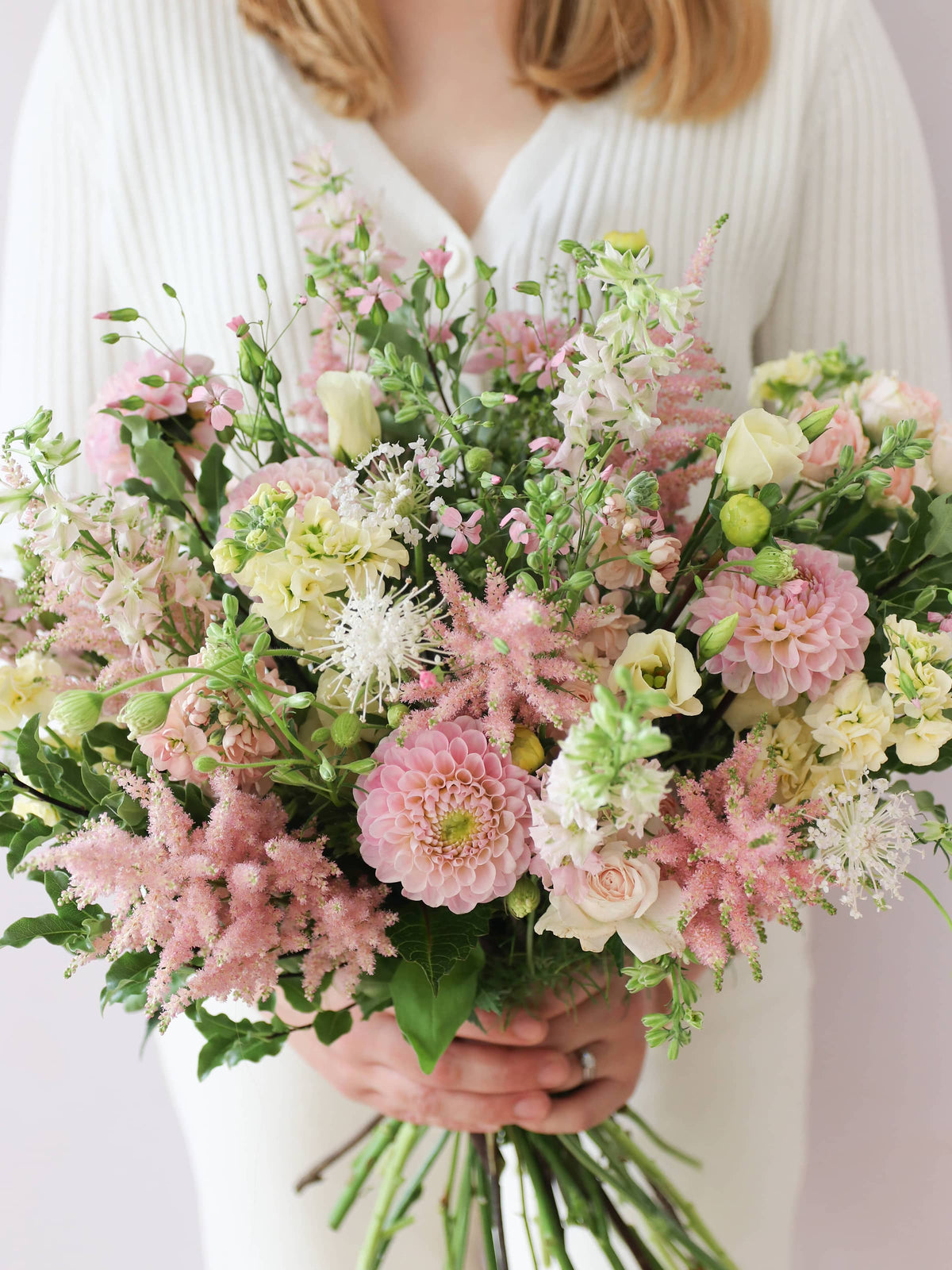 A woman wearing a white jumper and cream jeans, holding a pastel yellow and pink bouquet of flowers including Astilbe, Dahlias, Roses and Lisianthus in both hands.
