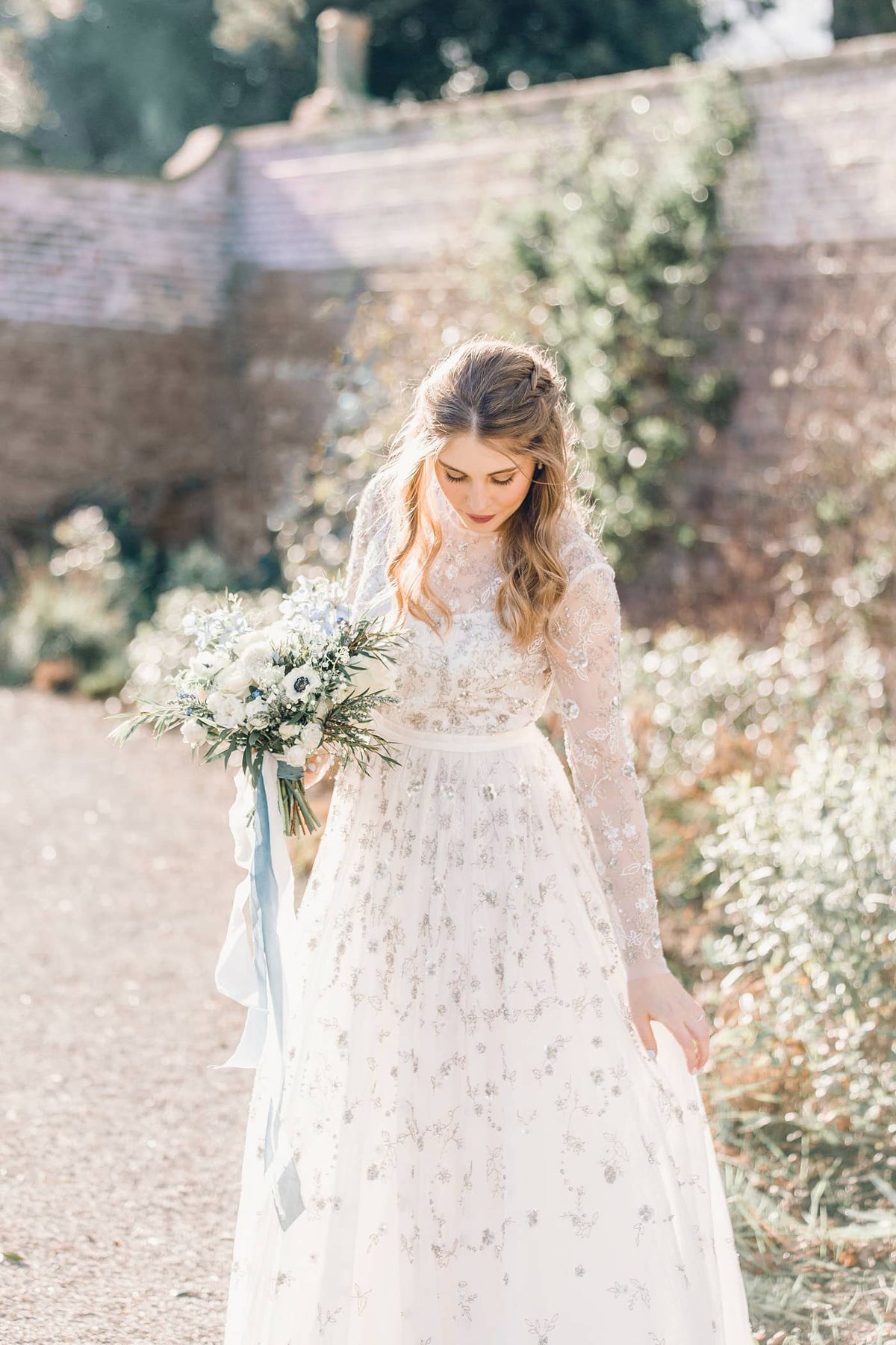 A woman in an embellished white bridal dress holding a bouquet of white and blue flowers with trailing blue ribbons on a sunny day in the gardens at Middleton Lodge, The Fig House.