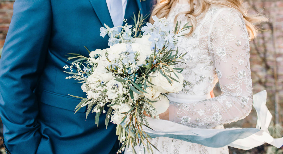 A groom in a blue suit and woman in an embellished white bridal dress holding a bouquet of white and blue flowers with trailing blue ribbons at Middleton Lodge, The Fig House standing against a brick wall.