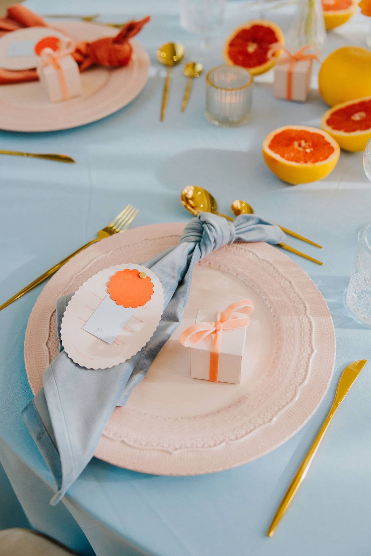 A wedding table setting with pastel pink charger plates, pastel pink favour boxes with a coral velvet ribbon and a blue. napkin, all sitting on a pastel blue tablecloth with gold cutlery.