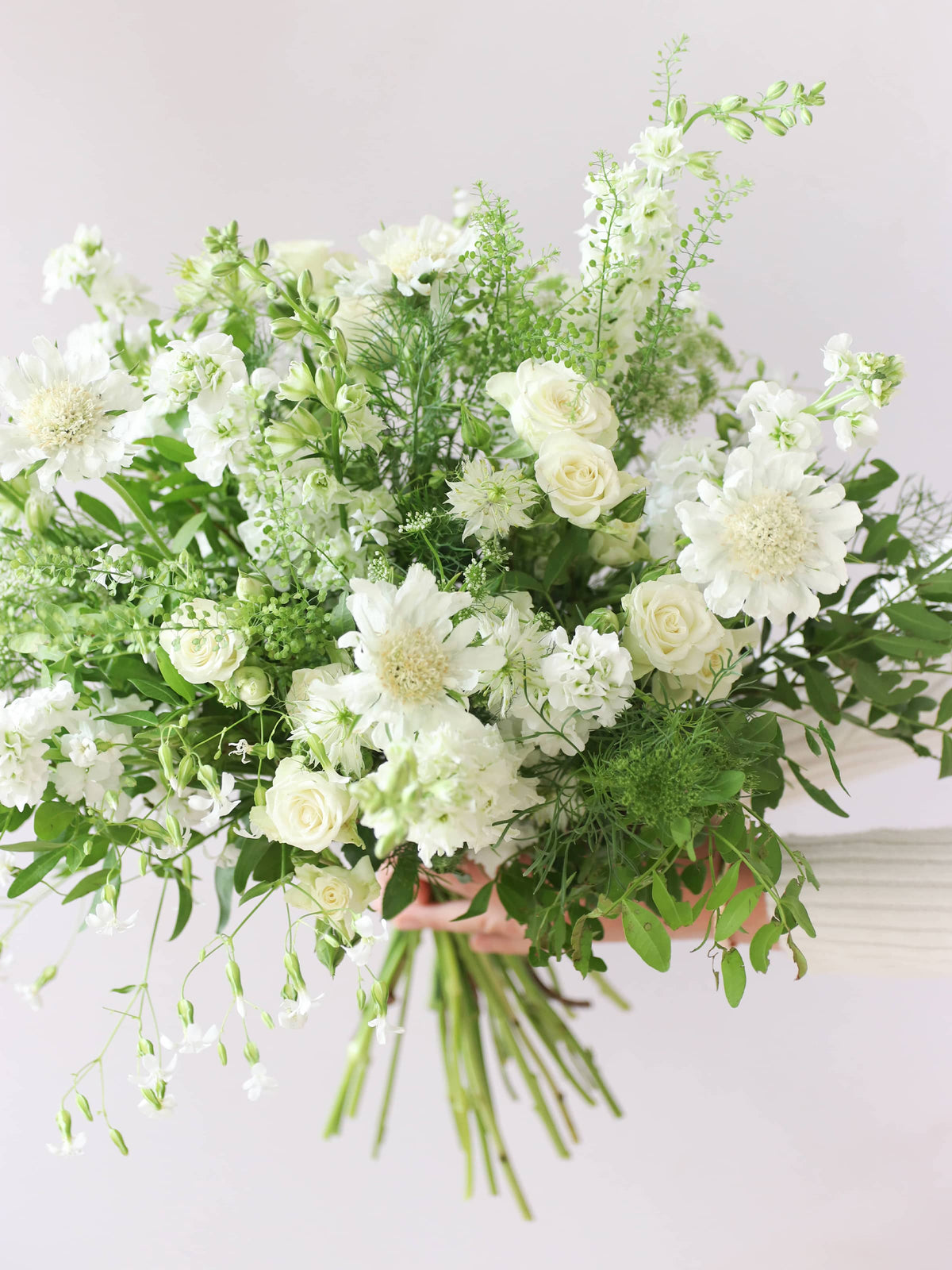 A woman holding a white and green bouquet of flowers out to the side against a neutral background. The bouquet includes Delphiniums, Stocks, Roses and Ammi.