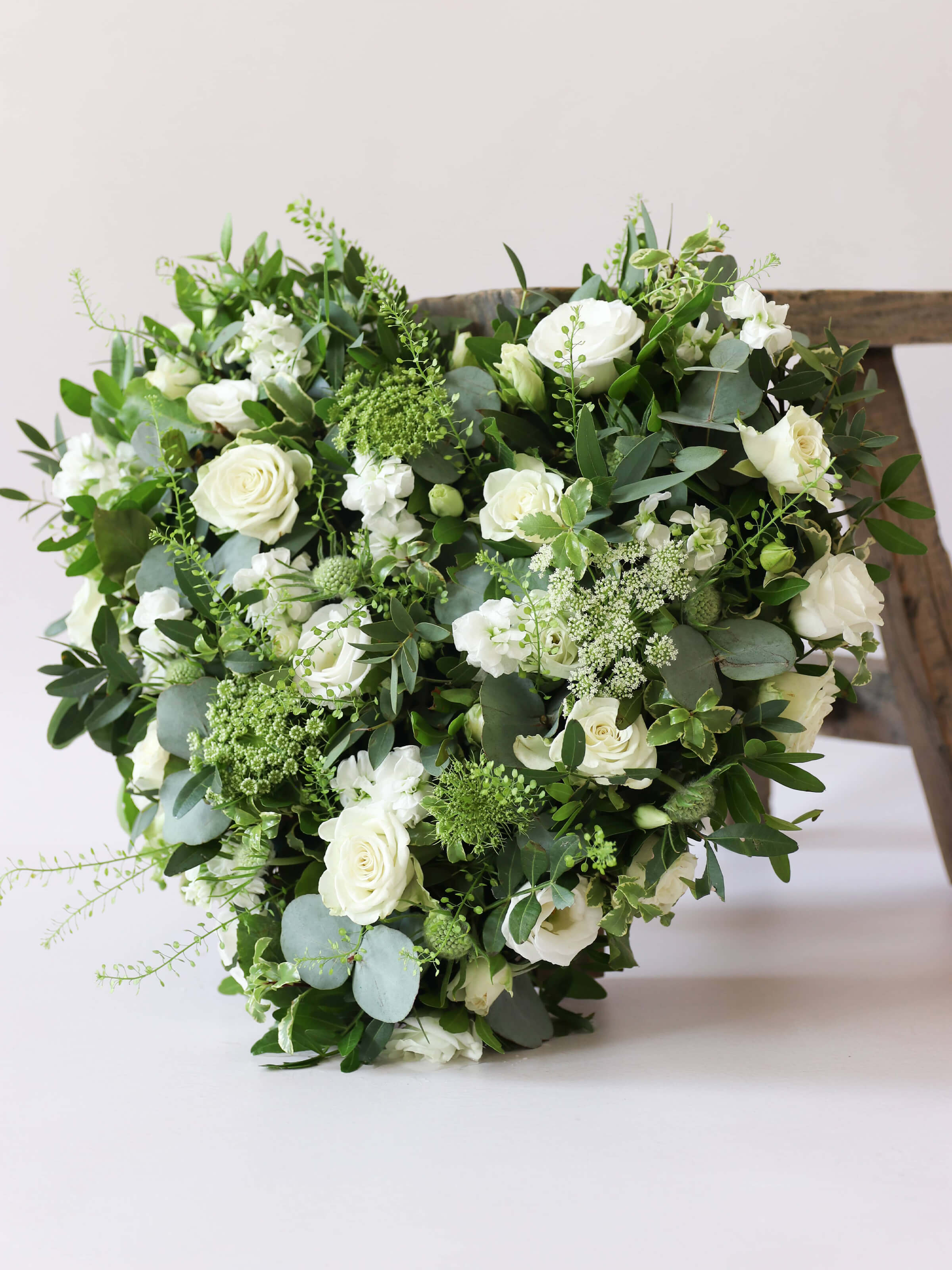 Heart shaped funeral posy with white and green flowers on a white background leaning against a wooden stool