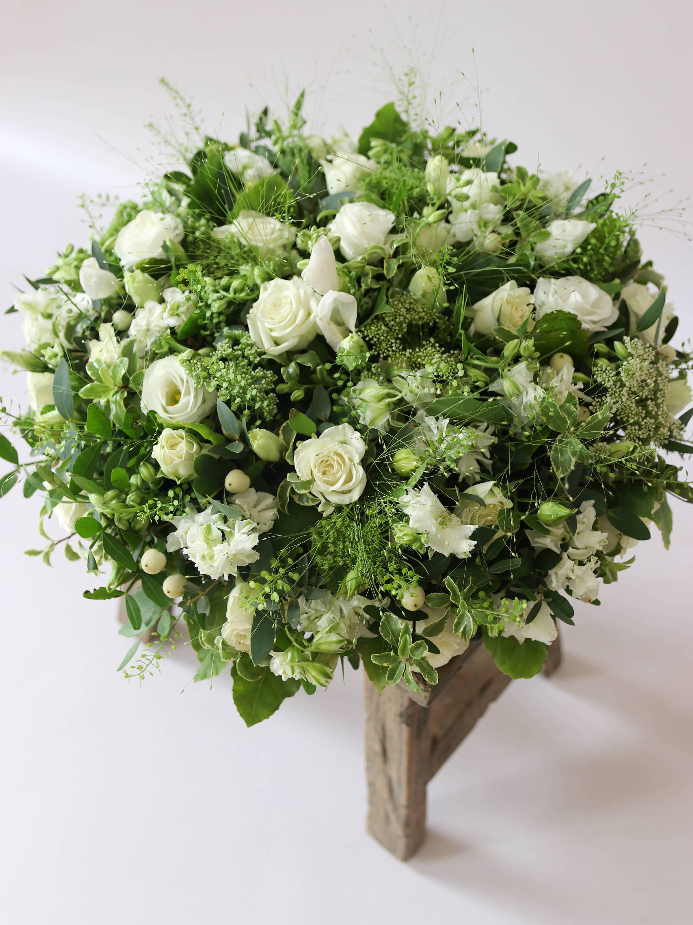 Round funeral posy with white and green flowers on a white background sitting on a wooden stool