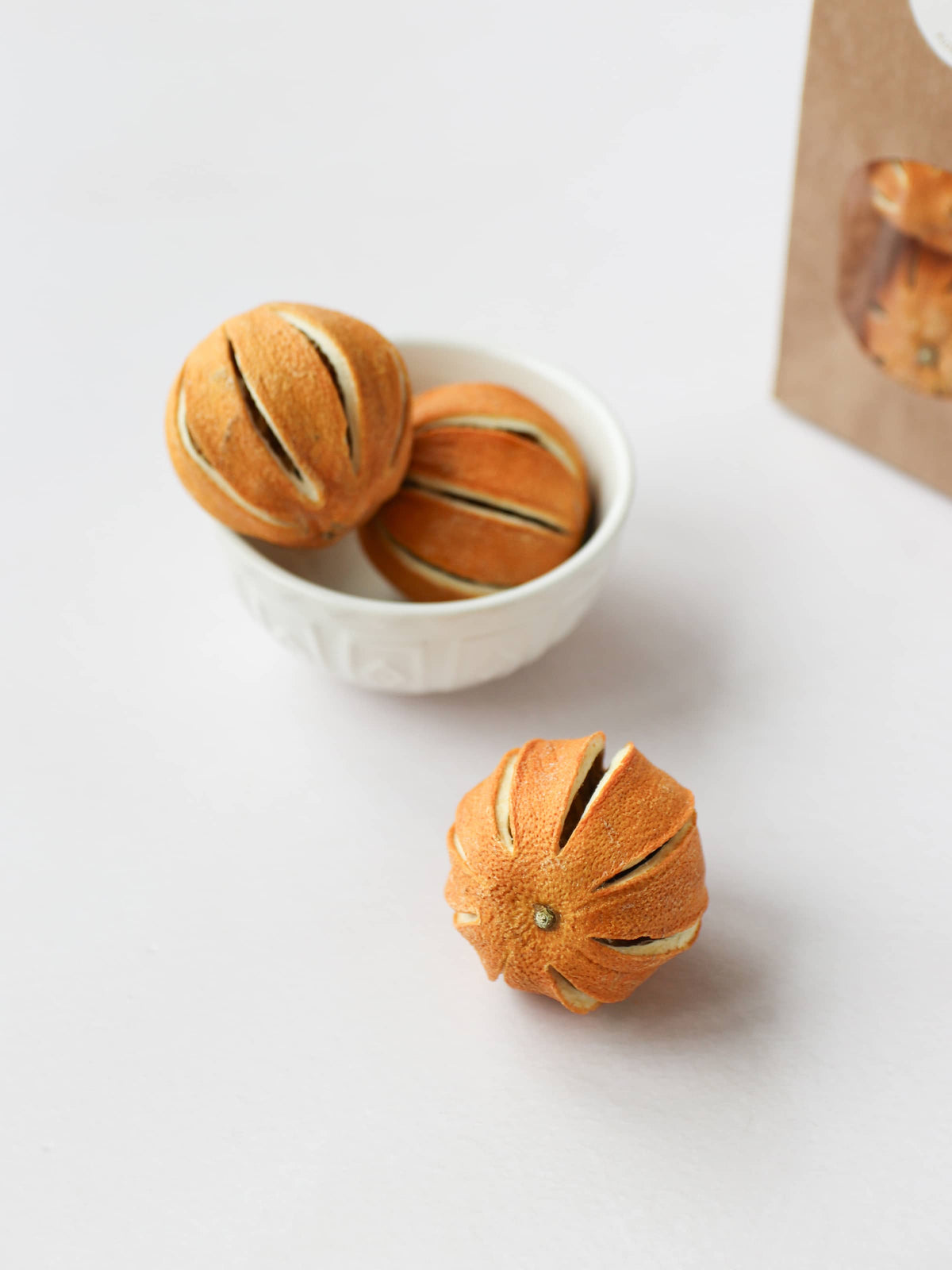 Three dried whole oranges for Christmas wreath making, positioned in a cream decorative bowl, on a light background. There is a bag of whole dried oranges in a brown kraft bag in the background.