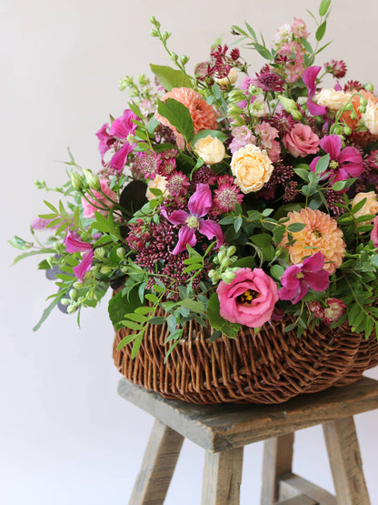 Close up angled arrangement of bright pink and peach flowers with green foliage in a wicker basket on a wooden stool against a light background