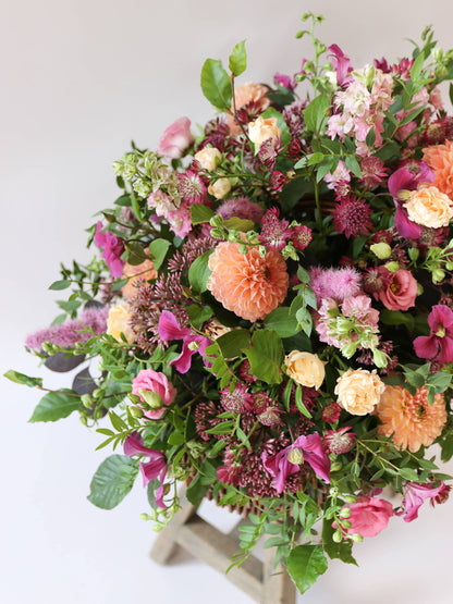 Top down view of bright pink and peach flowers with green foliage in a wicker basket on a wooden stool against a light background