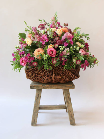 Arrangement of bright pink and peach flowers with green foliage in a wicker basket on a wooden stool against a light background