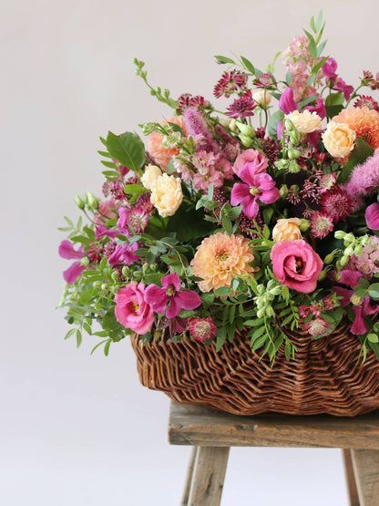 Close up side angle of a wicker basket with an arrangement of bright pink and peach flowers with green foliage in a wicker basket on a wooden stool against a light background