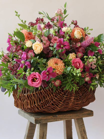 Wicker basket with an arrangement of bright pink and peach flowers with green foliage in a wicker basket on a wooden stool against a light background