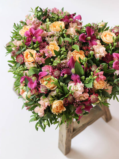 Heart-shaped posy with bright pink and peach flowers on a white background, sitting atop a wooden stool.