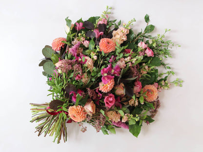 Top down view of a whole tied funeral sheaf with flowers including pink Lisianthus, peach Stocks, pink Delphiniums and coral Dahlias, tied with a burgundy satin bow.