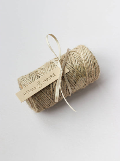 A spool of brown kraft mossing twine tied with a cream satin ribbon and a Petals & Paperie gift tag, sitting on a neutral background.