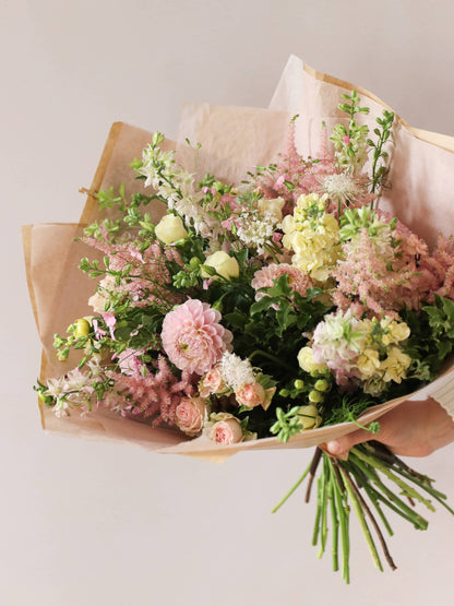 A person holding a pastel yellow and pink gift-wrapped bouquet of flowers including Astilbe, Dahlias, Roses and Lisianthus in one hand against a neutral background.