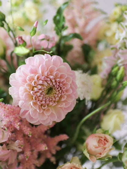 Close-up photograph of a pink Dahlia in a pastel pink and yellow bouquet, surrounded by other flowers and greenery