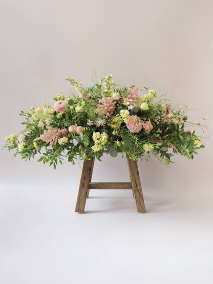 A side view of a 3ft coffin spray featuring pastel pink, yellow and peach flowers and foliage. The funeral arrangement is sitting on a rustic wooden stool against a neutral background and features Roses, Astlibe, Stocks and Lisianthus. 
