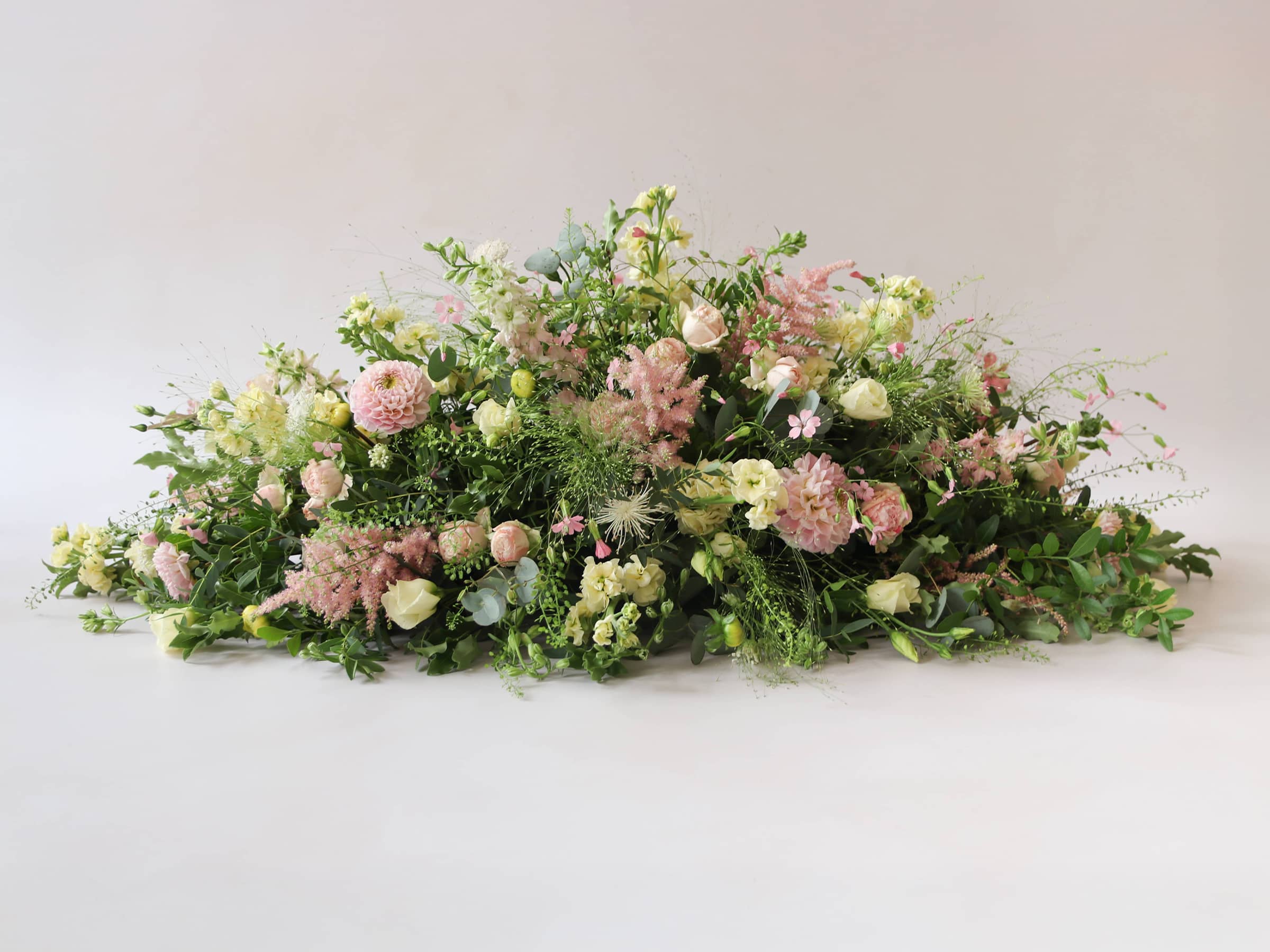 A side view of a 3ft coffin spray featuring pastel pink, yellow and peach flowers and foliage. The funeral arrangement is sitting against a neutral background and features Roses, Astlibe, Stocks and Lisianthus. 