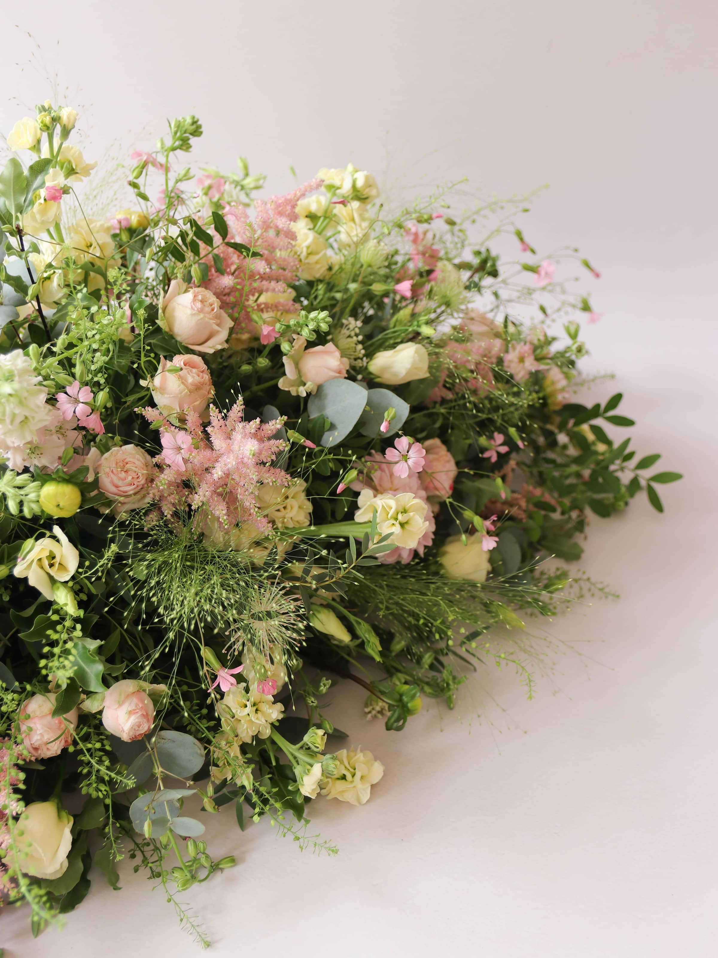 A side view of a 3ft coffin spray featuring pastel pink, yellow and peach flowers and foliage. The funeral arrangement is sitting against a neutral background and features Roses, Astlibe, Stocks and Lisianthus. 
