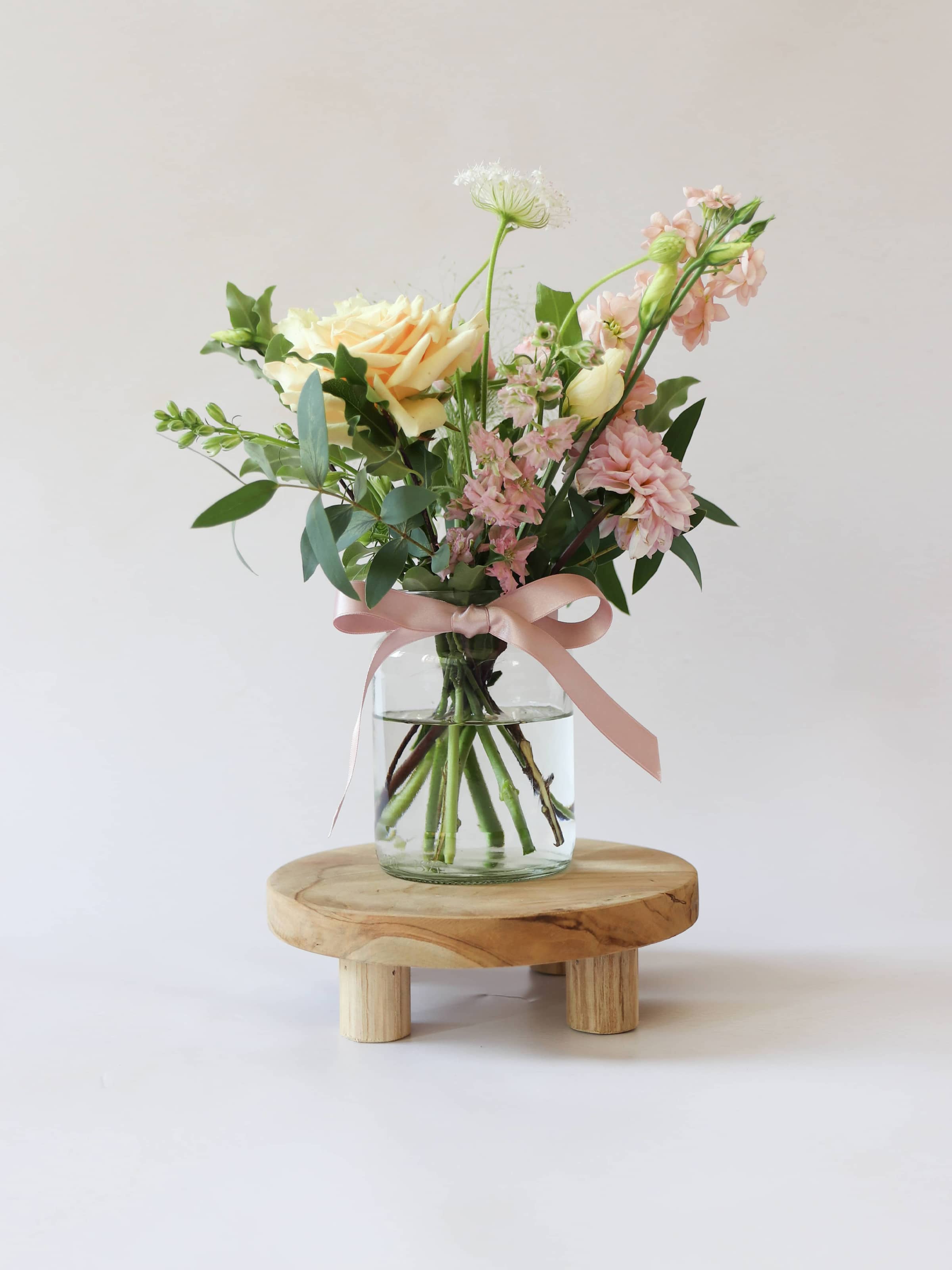 A single jam jar with pastel coloured flowers sitting on a wooden stool against a light background