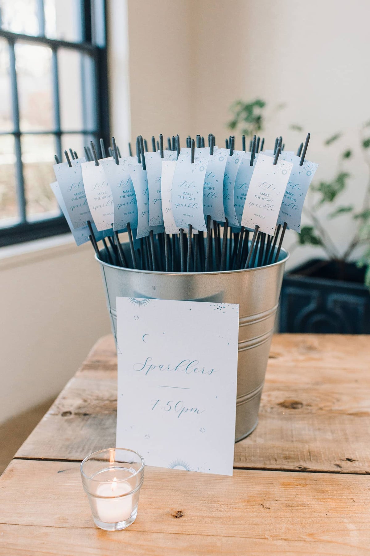 A metal bucket filled with giant sparklers with tags that read 'let's make the night sparkle' on a wooden banquet table with a candle and sign that reads 'sparklers at 7.50pm'.
