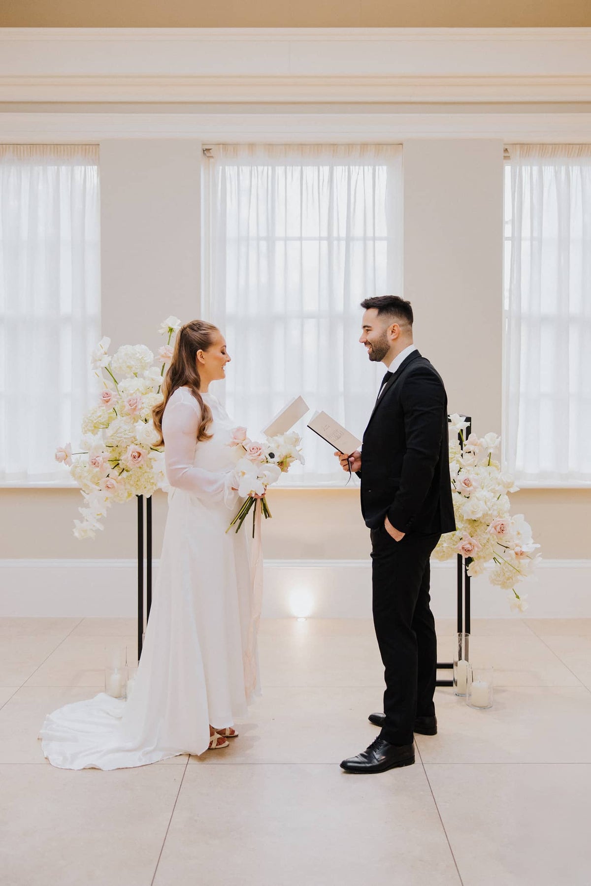 A groom in a black suit and a bride in a silk wedding dress, both holding a wedding vow book, tied with a black velvet ribbon and featuring a line illustration of Saltmarshe Hall on the front. They are standing in front of a white and blush floral installation.