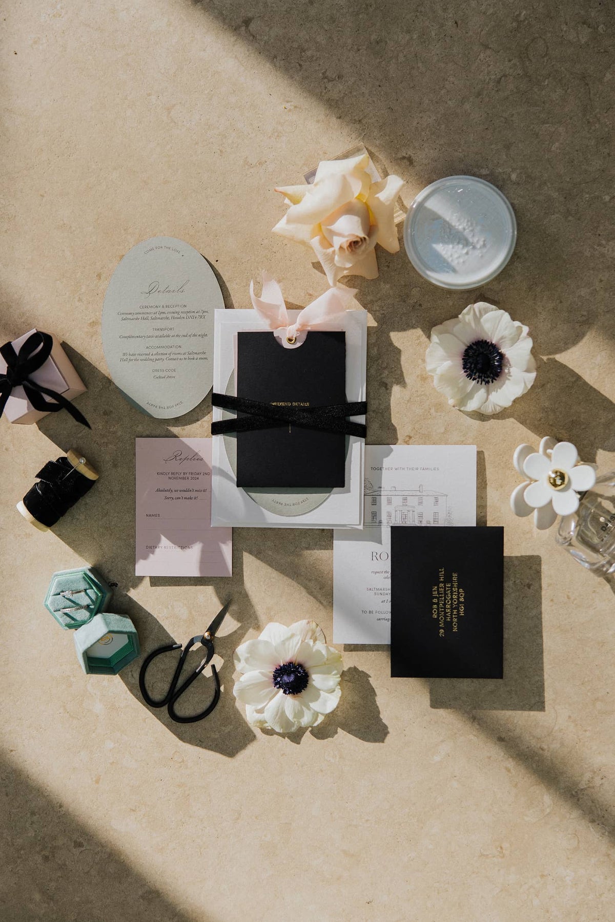 A flatlay of a wedding invitation suite, that features a line illustration of Saltmarshe Hall, gold foil and black and blush pink colours, lying on a textured surface.