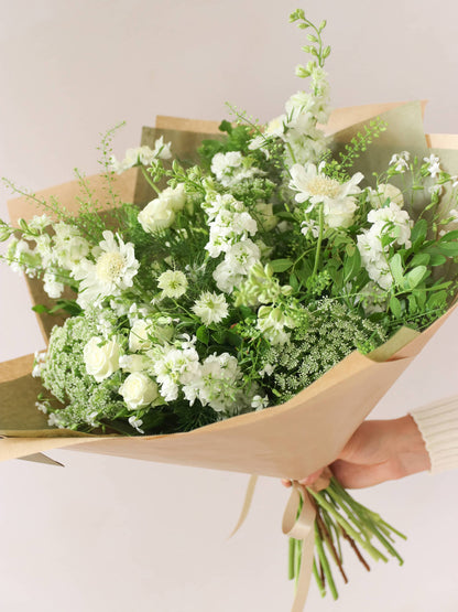 A woman holding a white and green gift-wrapped bouquet of flowers out to the side against a neutral background. The bouquet includes Delphiniums, Stocks, Roses and Ammi.