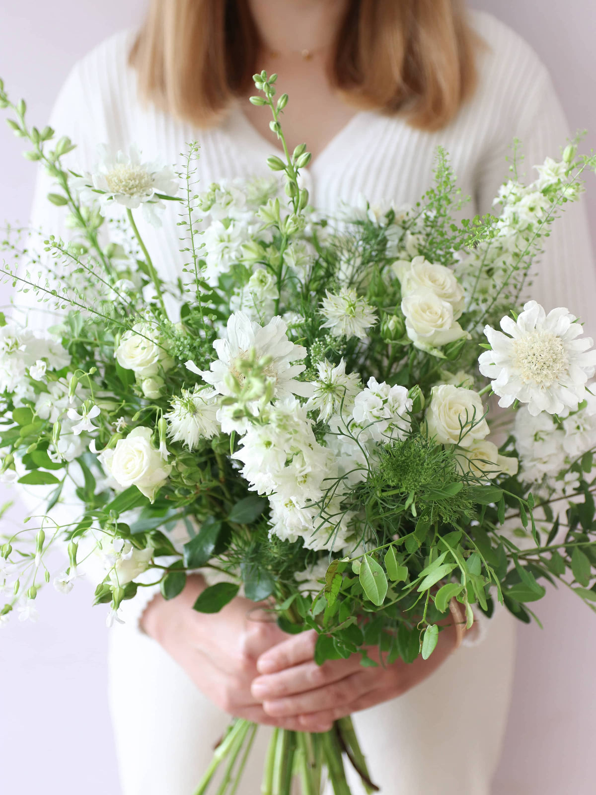 A woman wearing a white jumper and cream jeans, holding a white and green bouquet of flowers including Delphiniums, Stocks, Roses and Ammi in both hands against a neutral background.