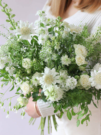 A woman wearing a white jumper and cream jeans, holding a white and green bouquet of flowers including Delphiniums, Stocks, Roses and Ammi in both hands against a neutral background.