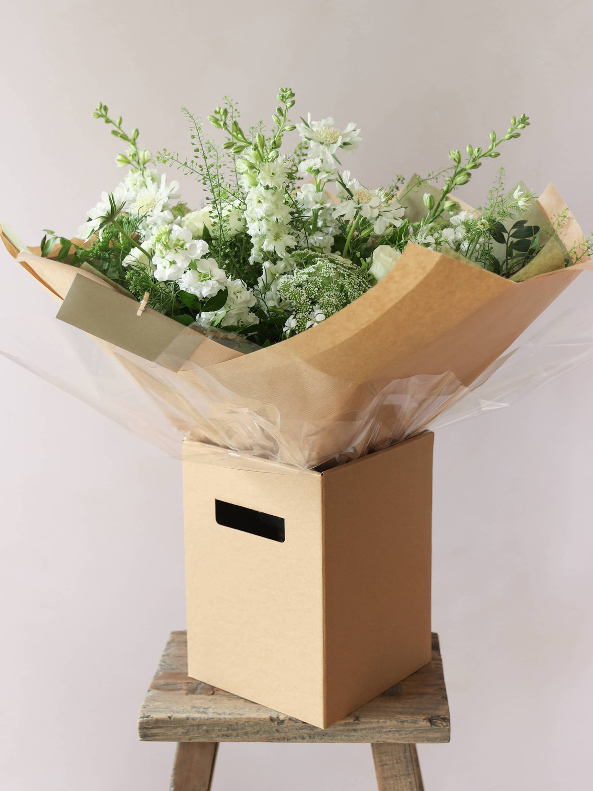 A gift-wrapped bouquet of green and white flowers standing in a kraft brown gift  box. The gift box is sitting on a small wooden stool against a neutral background.