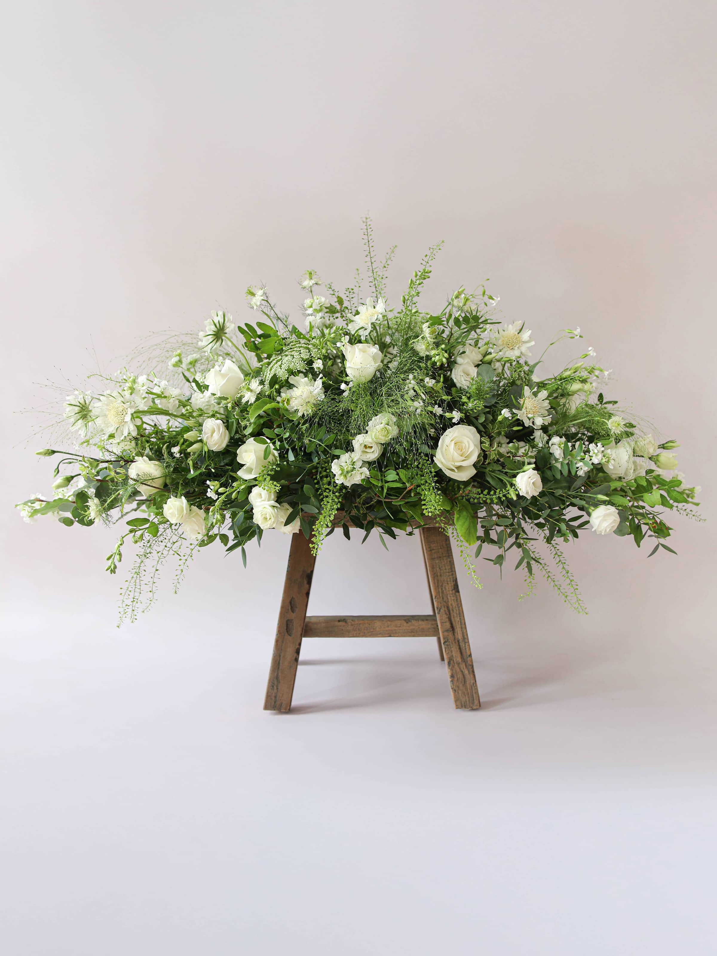 A side view of a 3ft coffin spray featuring white and green flowers and foliage. The funeral arrangement is sitting on a rustic wooden stool against a neutral background and features white Roses, Ammi, Stocks and Lisianthus. 