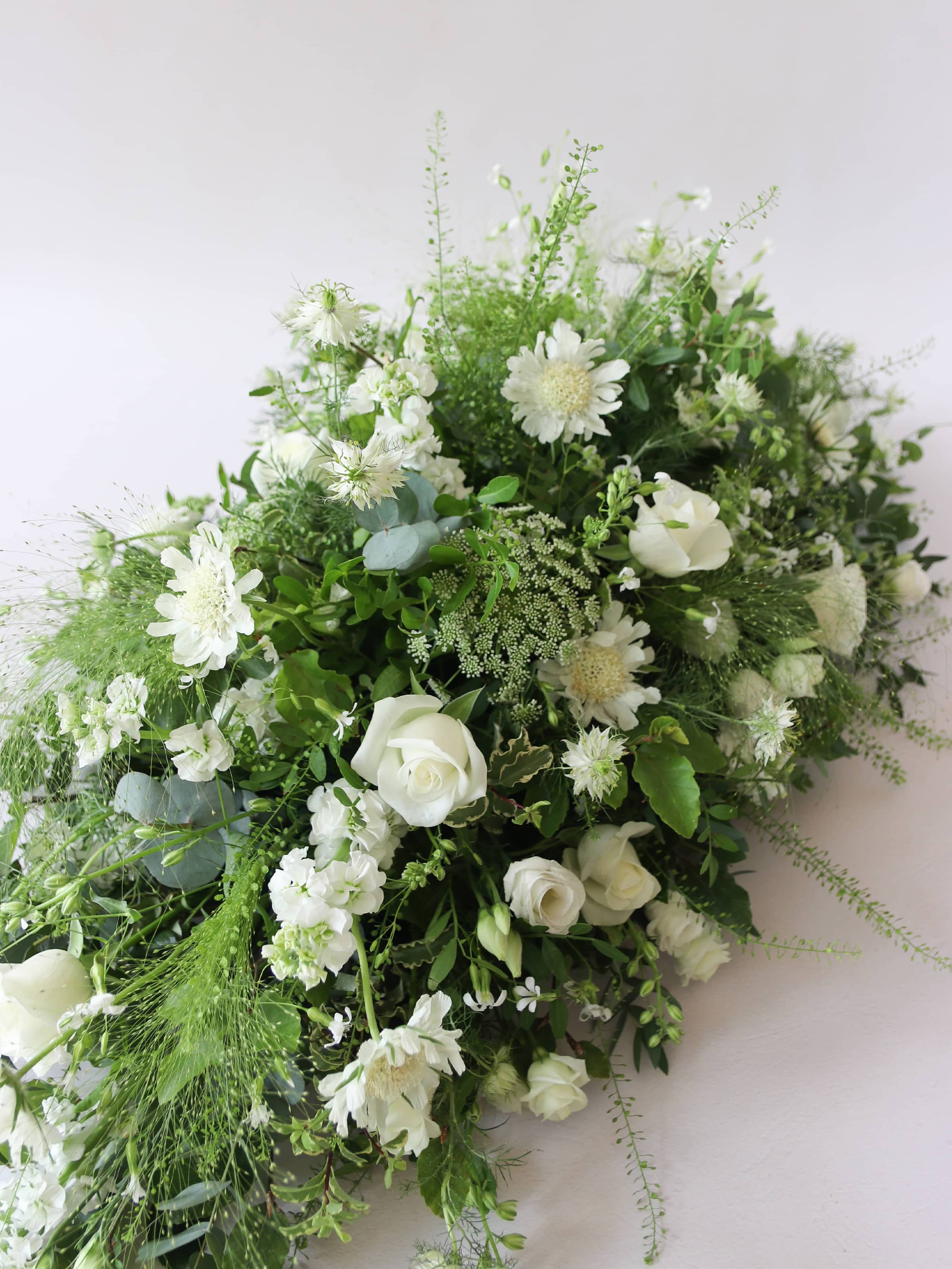 A diagonal top view of a 3ft coffin spray featuring white and green flowers and foliage. The funeral arrangement is sitting against a neutral background and features white Roses, Ammi, Stocks and Lisianthus. 